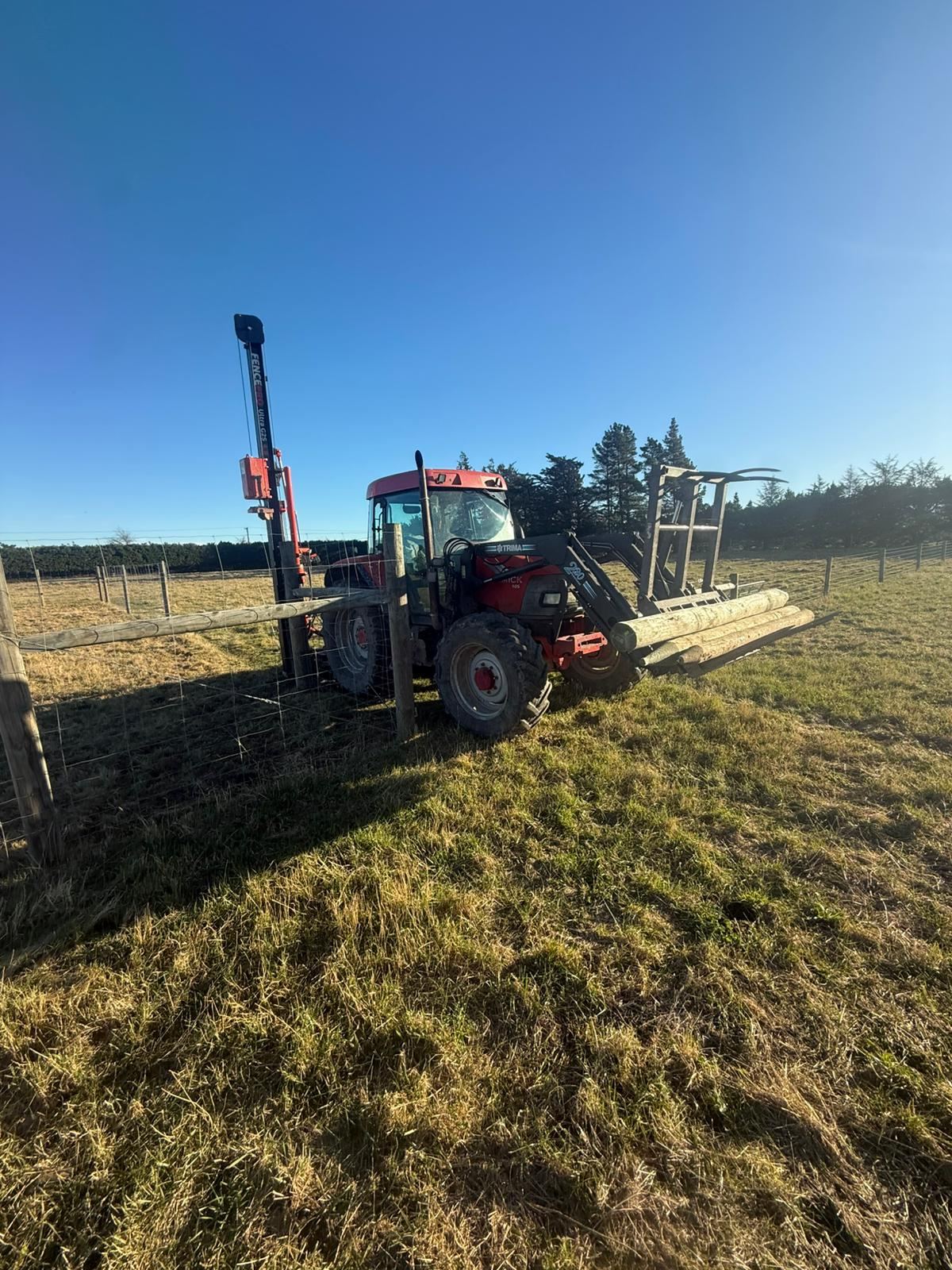 A tractor-mounted post driver working alongside a tall deer fence of netting and timber posts