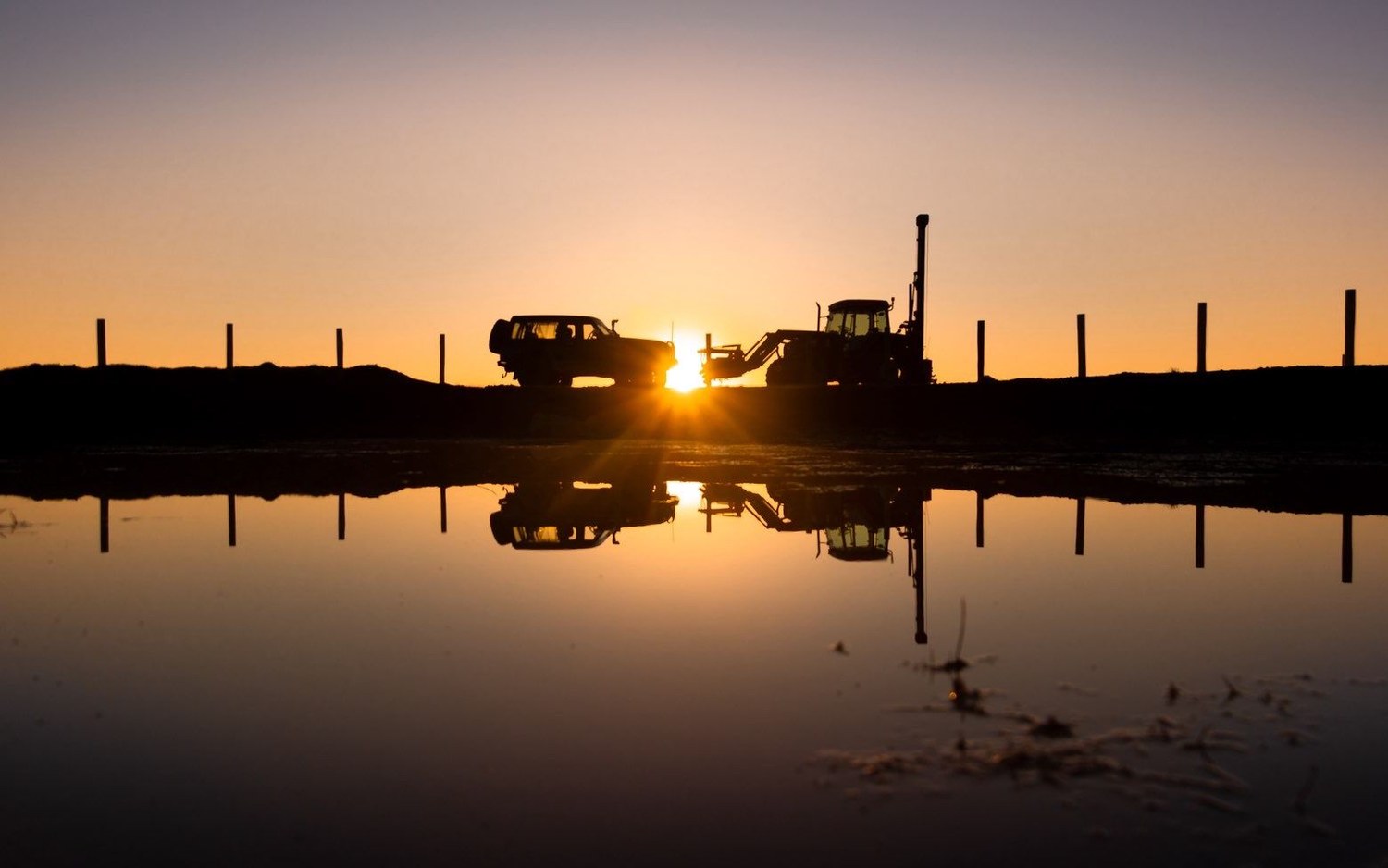 A new fence line at dusk, posts running to the horizon on both sides with the ute and post driver silhouetted against the setting sun and mirrored in still water.