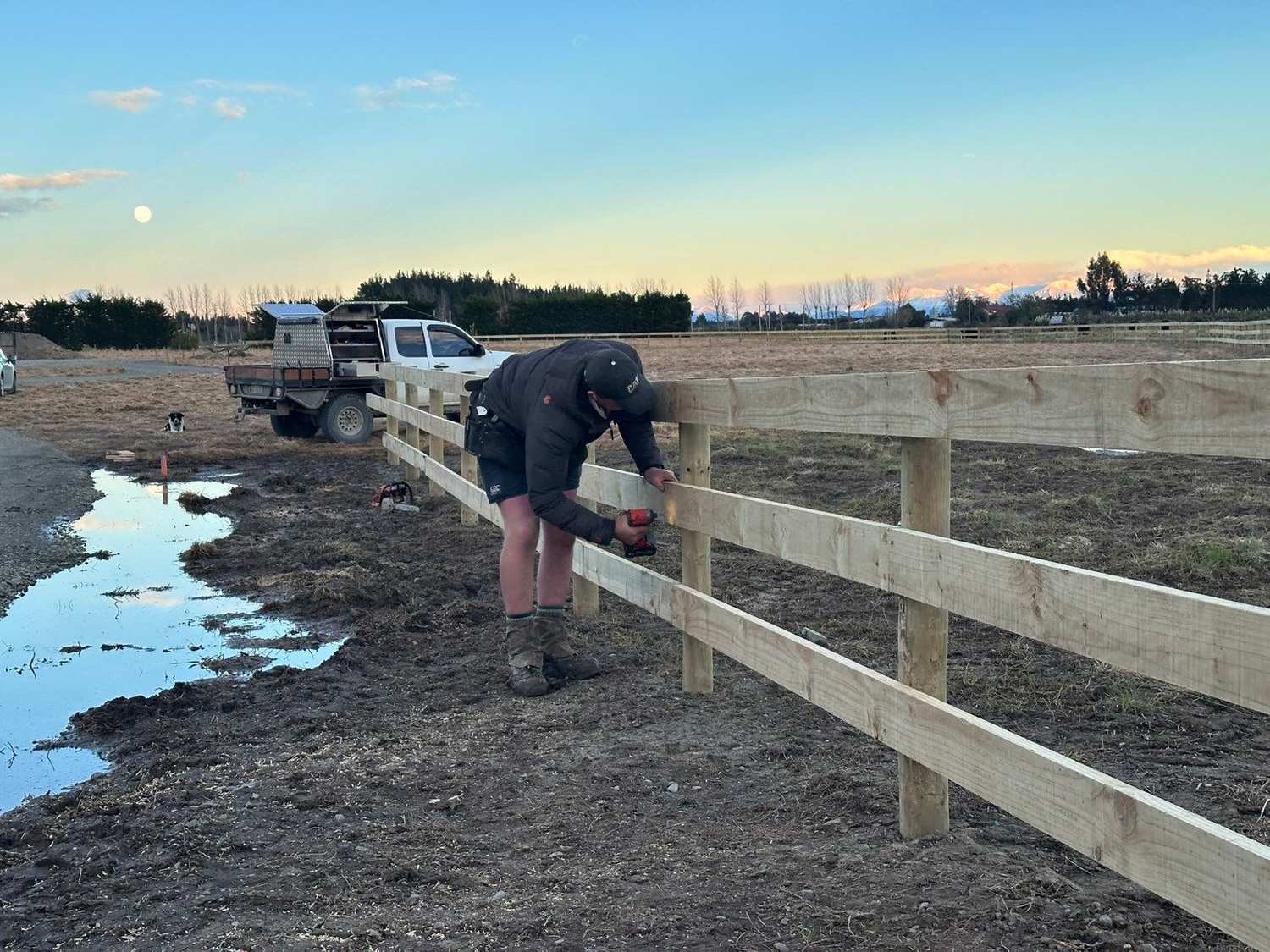Nathan fixing a rail on a new post and rail fence, ute and dog behind him and the Southern Alps on the horizon